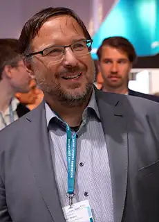 Ein Mann mit Bart, Brille und Anzug lächelt an einem Messestand.